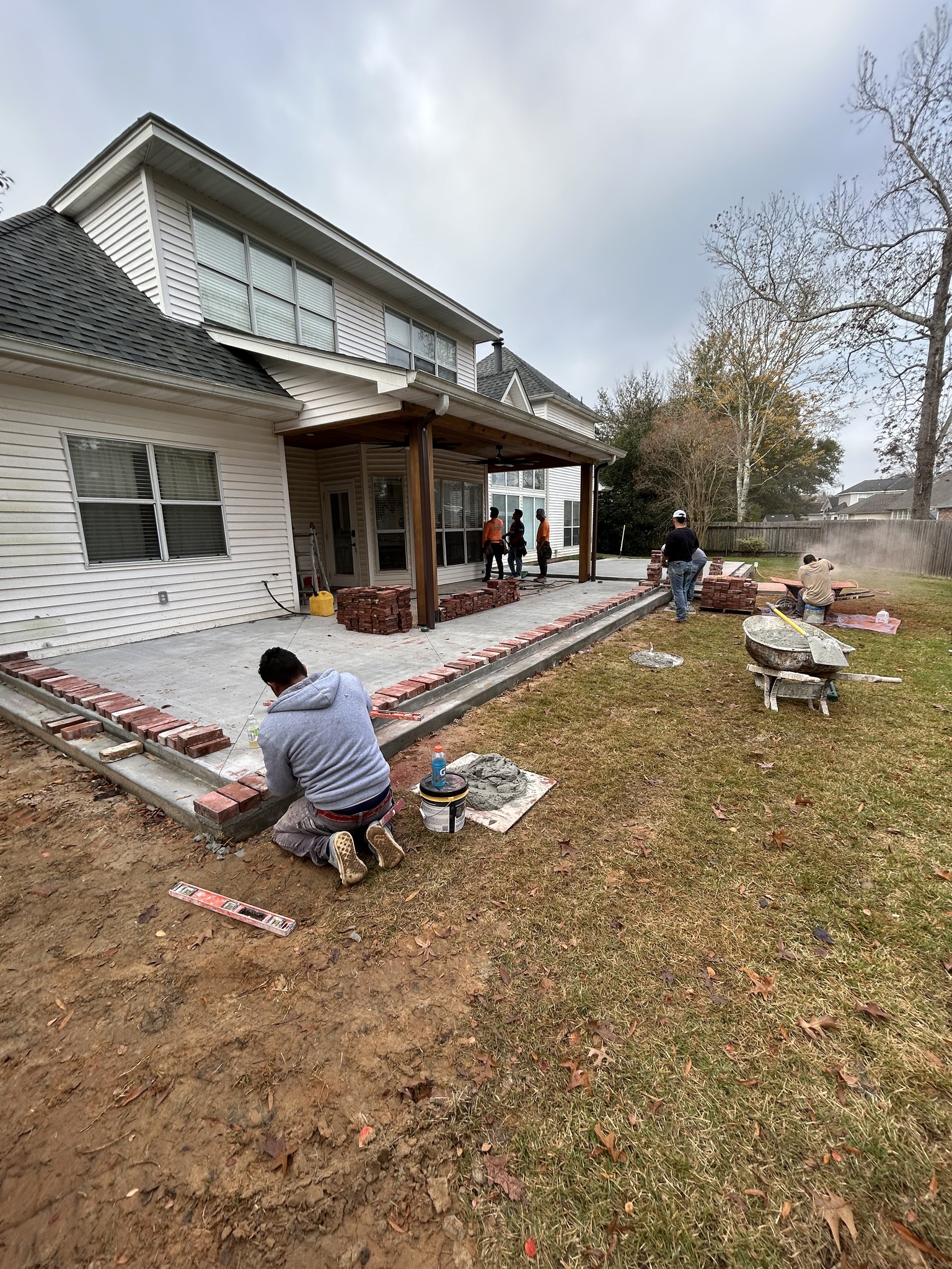 Several people work on constructing a brick patio in the backyard of a white two-story house, with tools and materials scattered around the area.