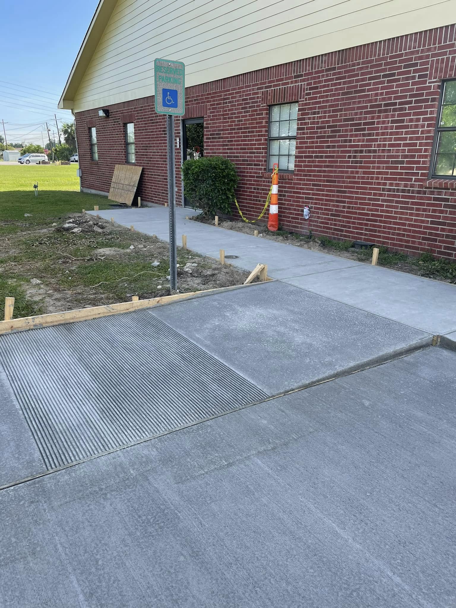 Freshly poured concrete sidewalk and ramp with a handicapped parking sign in front of a red brick building; construction materials and a traffic cone are nearby.
