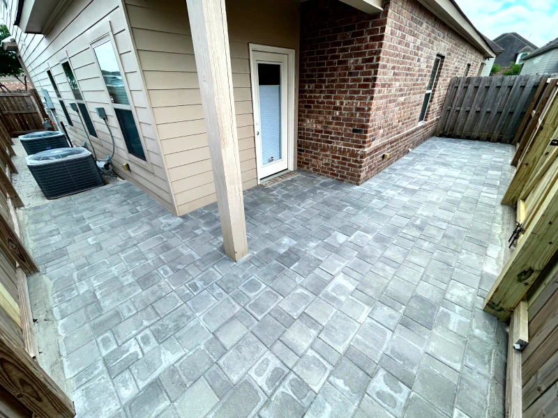 A fenced backyard patio with light gray stone pavers, beige siding, red brick walls, and two outdoor air conditioning units.