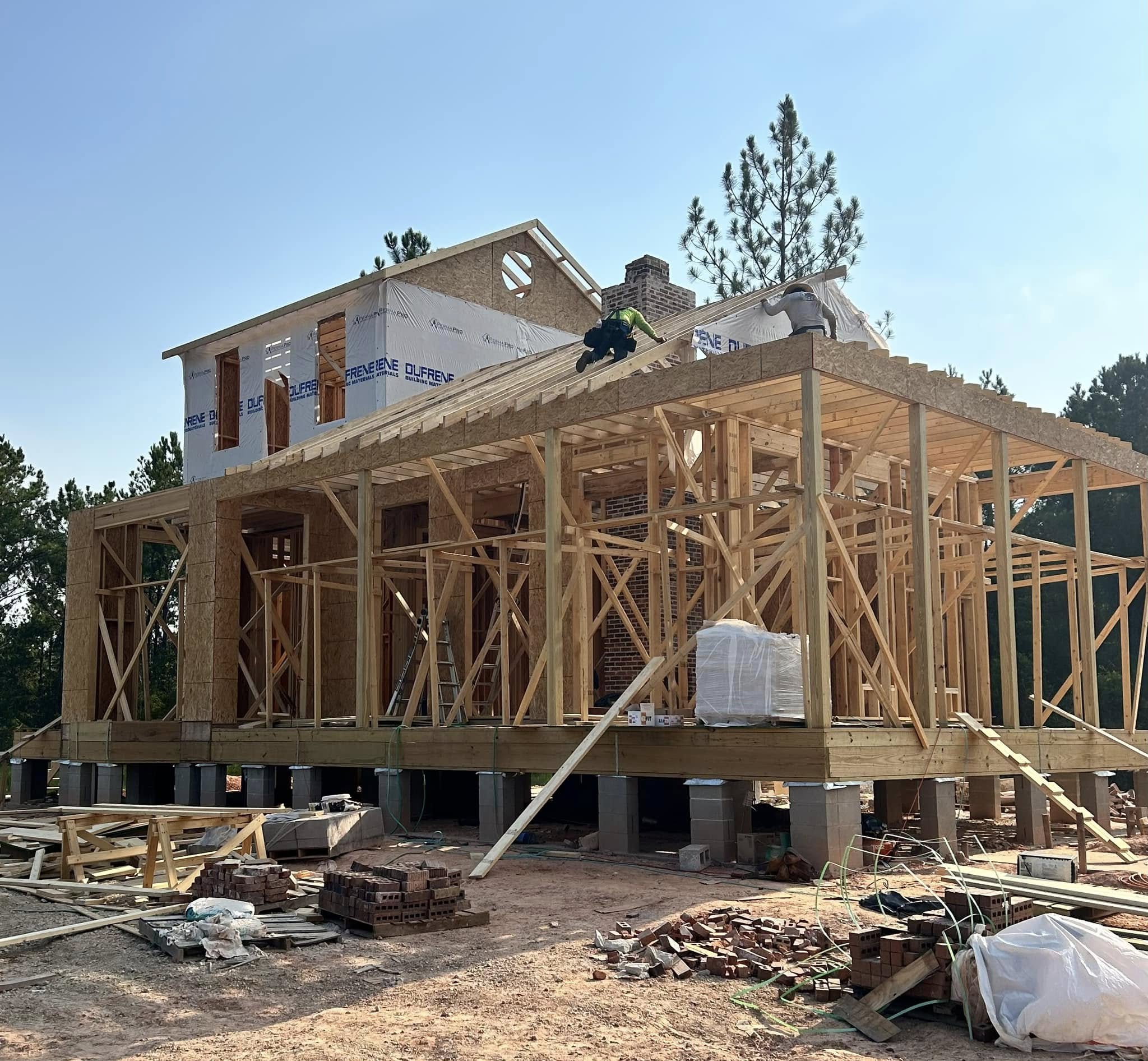 A two-story house under construction, showing exposed wooden framing, building materials on site, and the second floor partially covered with house wrap.