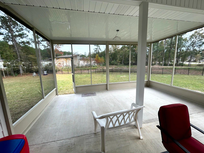 A screened-in porch with a white bench, a red chair, and an open view of a grassy backyard and trees outside.