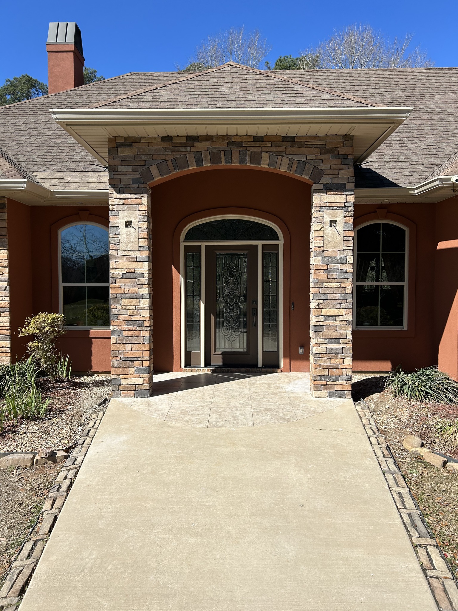 Front entrance of a house with stone columns, arched doorway, double glass doors, and two large windows on either side. Concrete path leads to the entry.