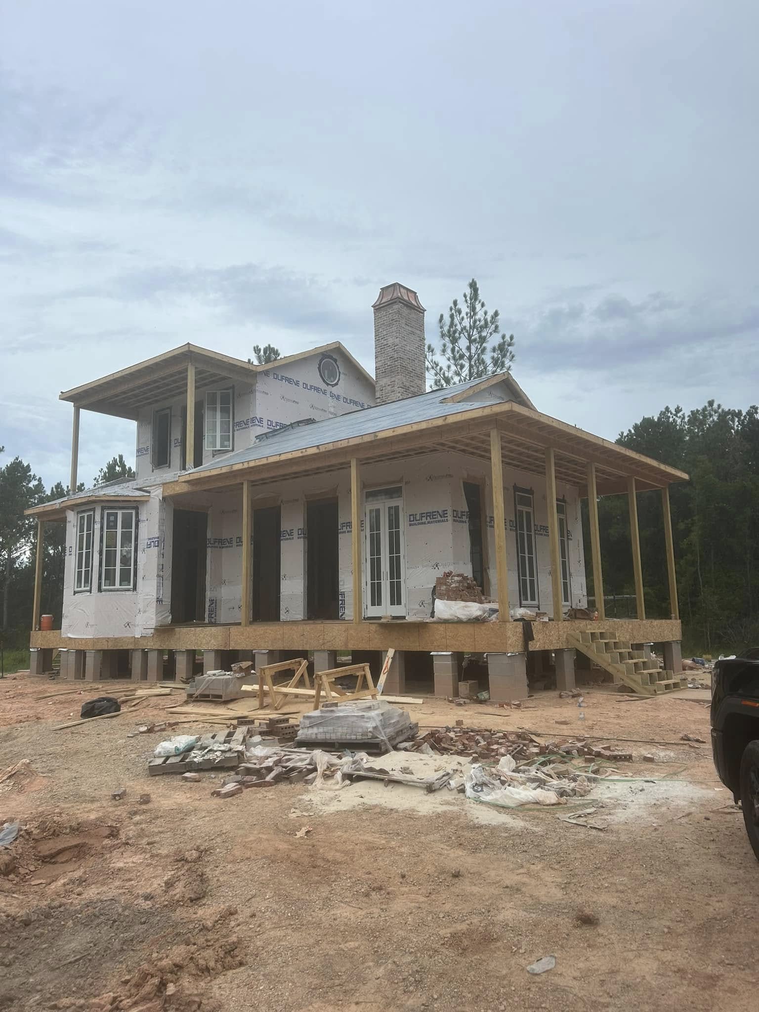 A partially constructed two-story house with a covered porch, surrounded by dirt, building materials, and trees in the background.