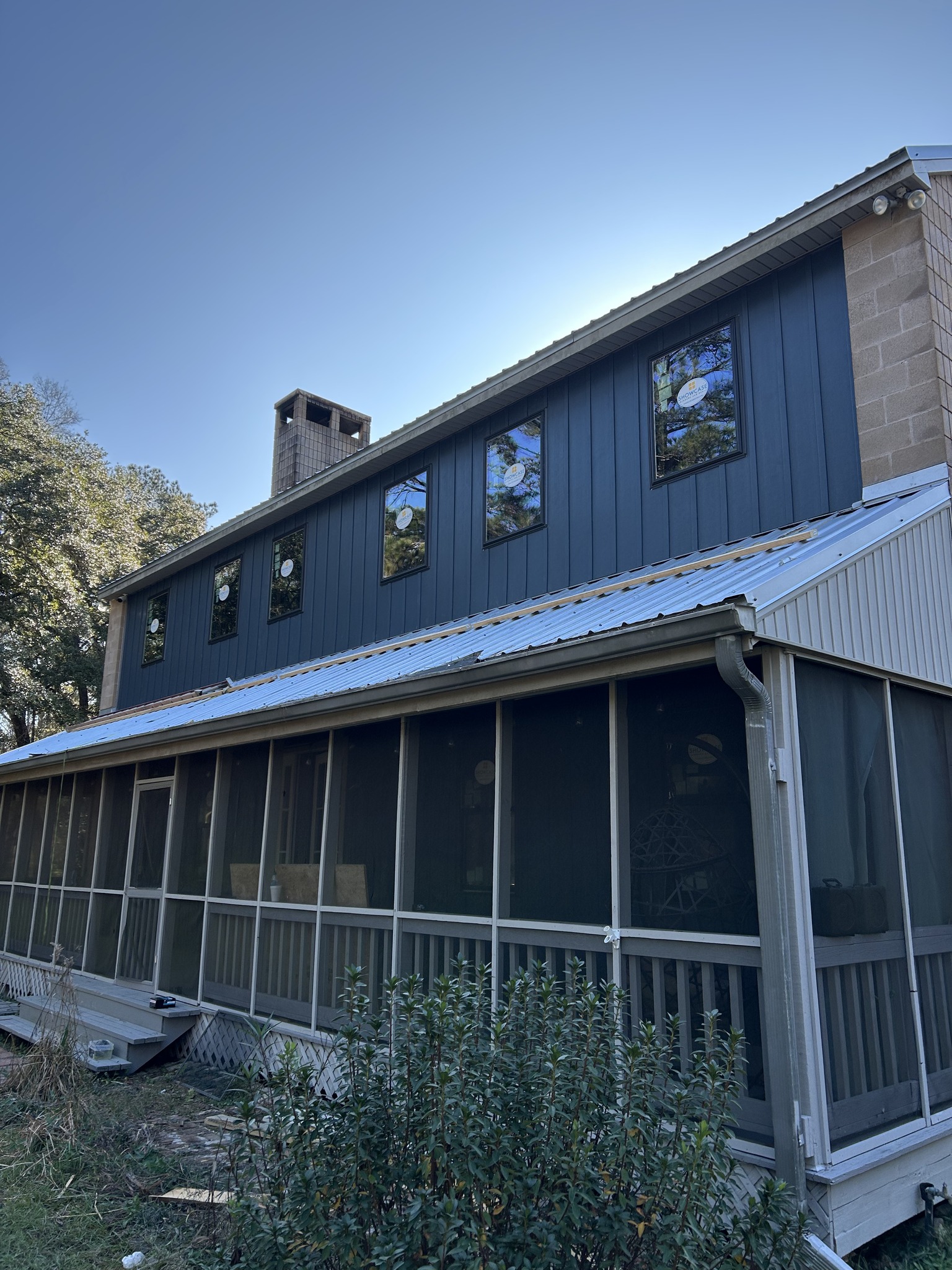 Two-story house with dark vertical siding, three upper windows, a brick chimney, and a screened porch on the lower level, set under a clear blue sky.